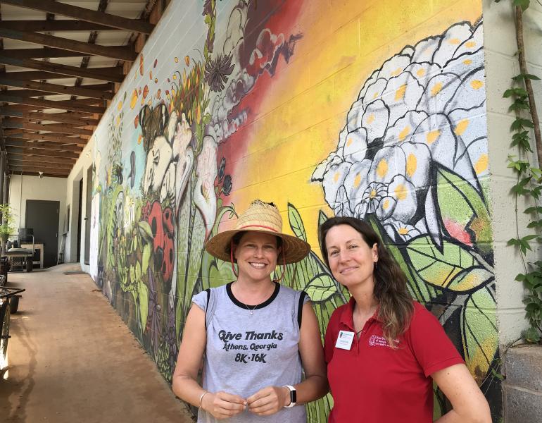 Two women in grey and red shirts stand in front of a colorful mural that shows flowers and insects at the State Botanical Garden of Georgia
