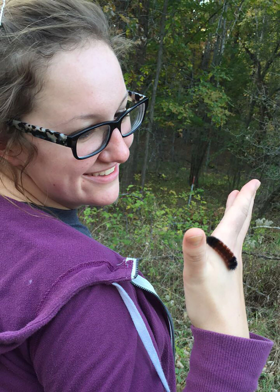 A white woman with glasses and wearing a purple hoodie, smiling at a large fuzzy caterpillar that is crawling over her right hand.
