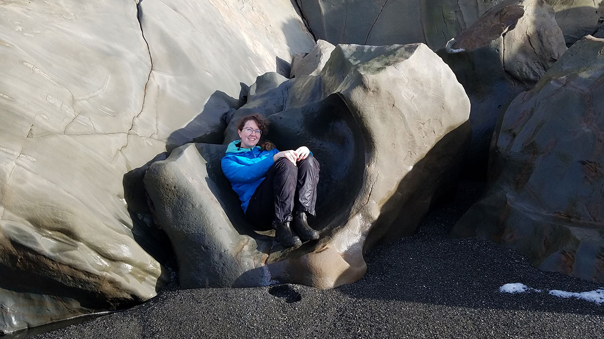 A white woman with curly hair, wearing waterproof hiking clothes, smiling and sitting on a very large rock that has been worn smooth from erosion. 