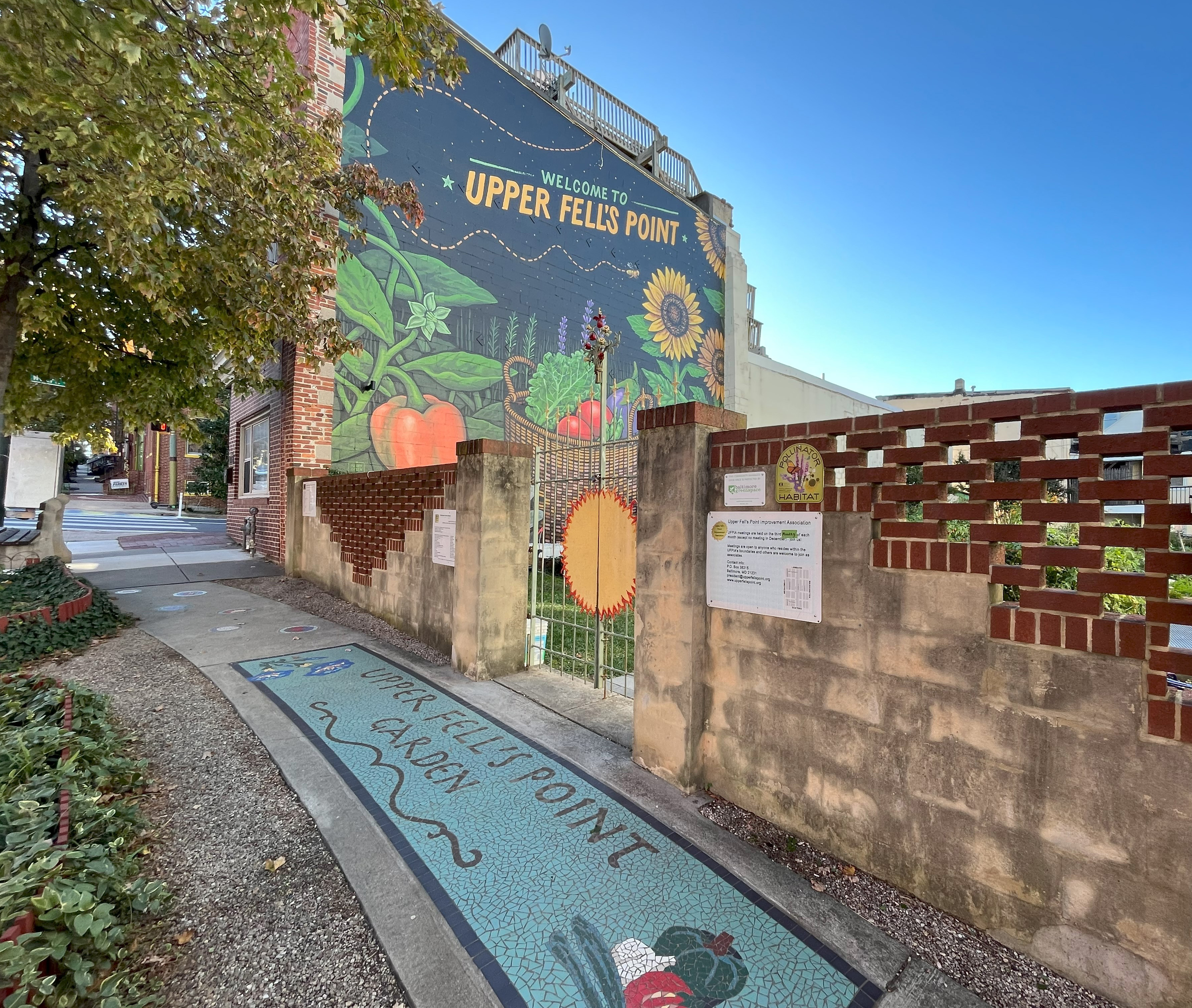 Giant mural featuring sunflowers, insects, and produce, above a brick garden fence and a mosaic sidewalk making the Upper Fell's Point garden