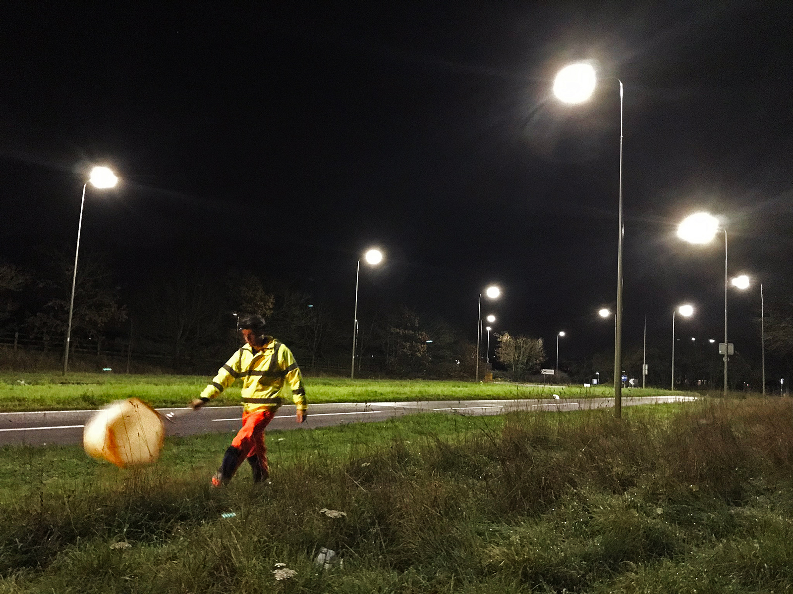 A person wearing fluorescent clothing and a headlamp swings a net alongside a roadway lit by streetlamps at night.