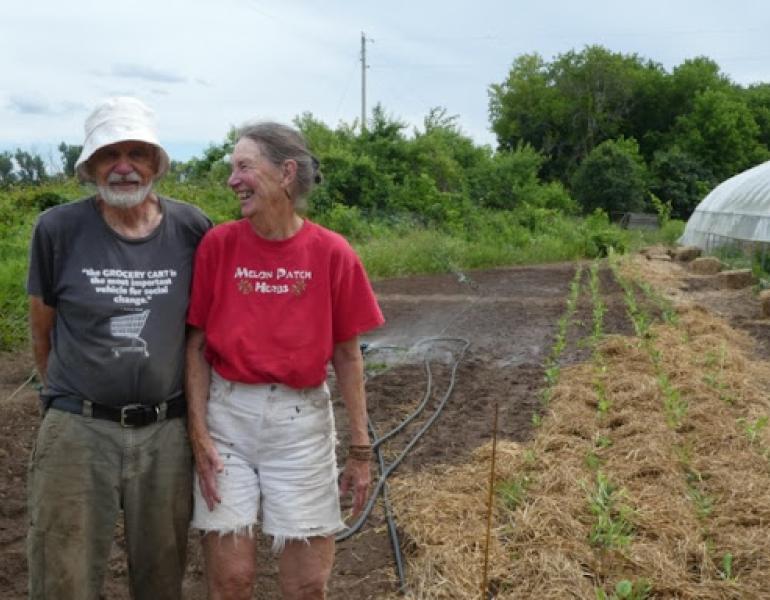 A laughing couple stands in front of several rows of planted seedlings