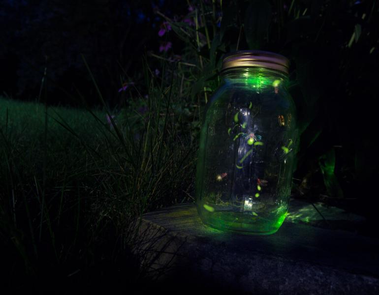 A glowing green mason jar contains a few fireflies in flight.