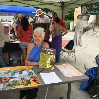  Phyllis Stiles, an older white woman with grey hair, presenting a table display at a local environmental festival. She is smiling and surrounded by information about pollinators. 